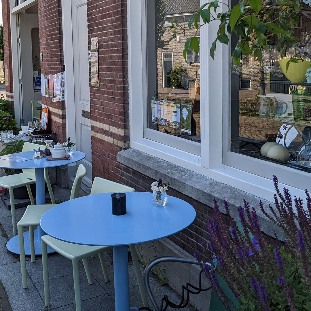 terras van Galerie Weerloos in Maasland langs de gevel van hun pand op een zomerse dag met twee blauwe tafeltjes en groene stoeltjes in Midden-Delfland