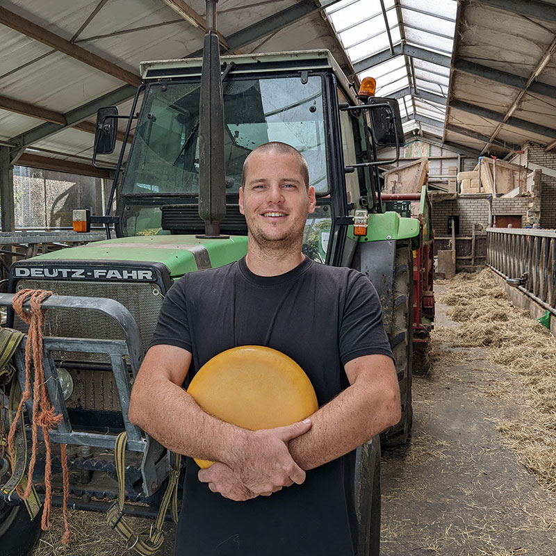 bas van den berg van Hoeve Ackerdijk in een stal met een tractor en een grote ronde gele kaas in zijn handen maker in midden-delfland boer