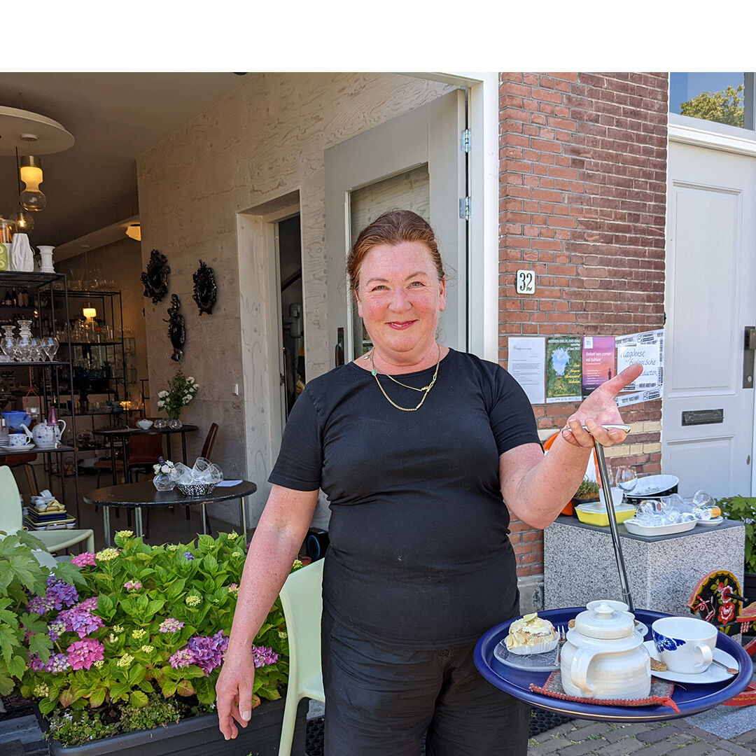 Marga van der Wulp voor het pand van Galerie Weerloos in Maasland in Midden-Delfland met een blad met thee en gebakjes gemaakt van streekproducten uit Midden-Delfland