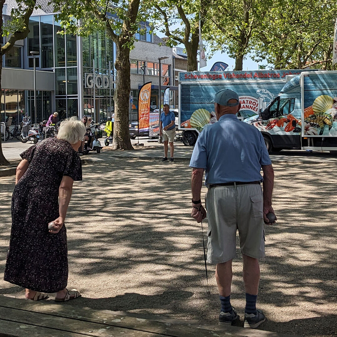 Koningin Julianaplein in Den Hoorn met petanque spelende ouderen onder platanen met groene bladeren en in de achtergrond kraampjes van een warenmarkt