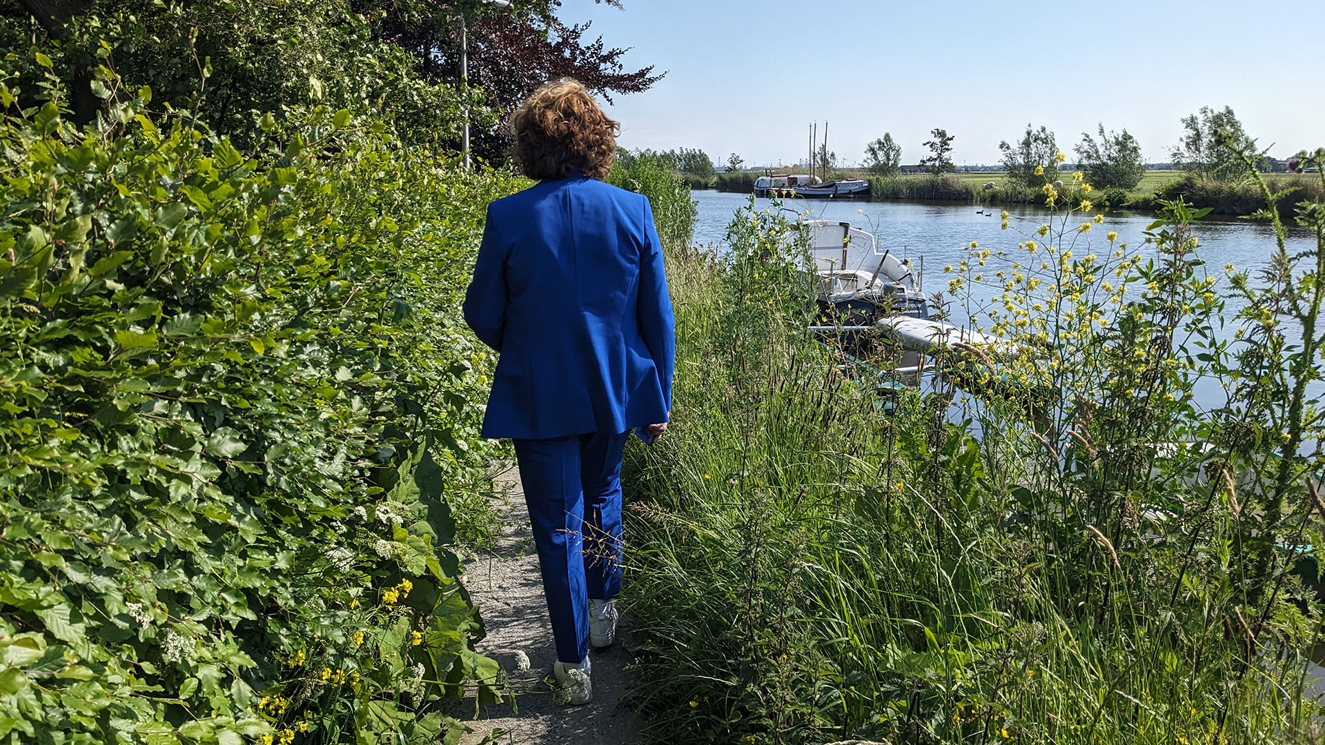 Marja van Bijsterveld, burgemeester van Delft wandelt langs de Vlaardingsevaart op een zonnige lentedag met links en rechts van het pad groene planten en bloemen en rechts het water met een boot